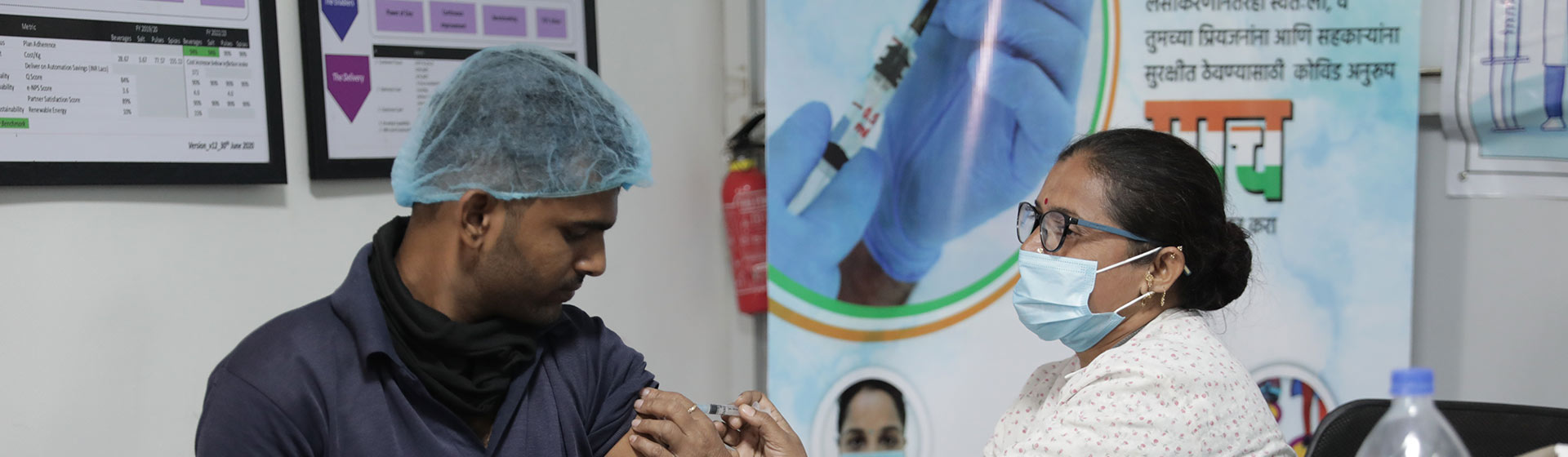 A nurse gives a vaccine shot to a man wearing a hair net, in a clinic promoting COVID-19 awareness and safety measures.